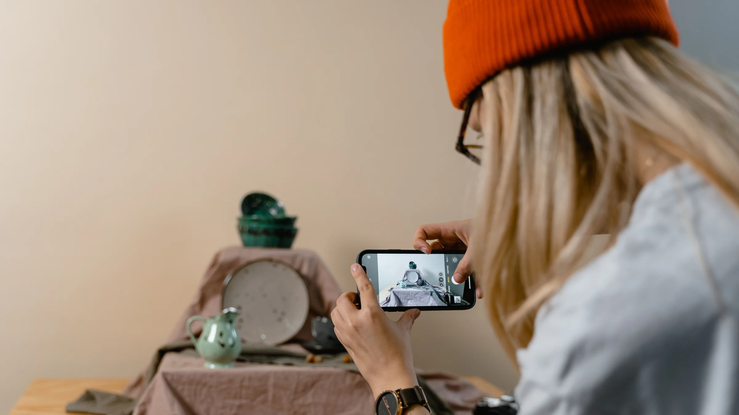 Person in an orange knit cap photographing a ceramic jug, plate and bowls on a cloth-covered table in daylight