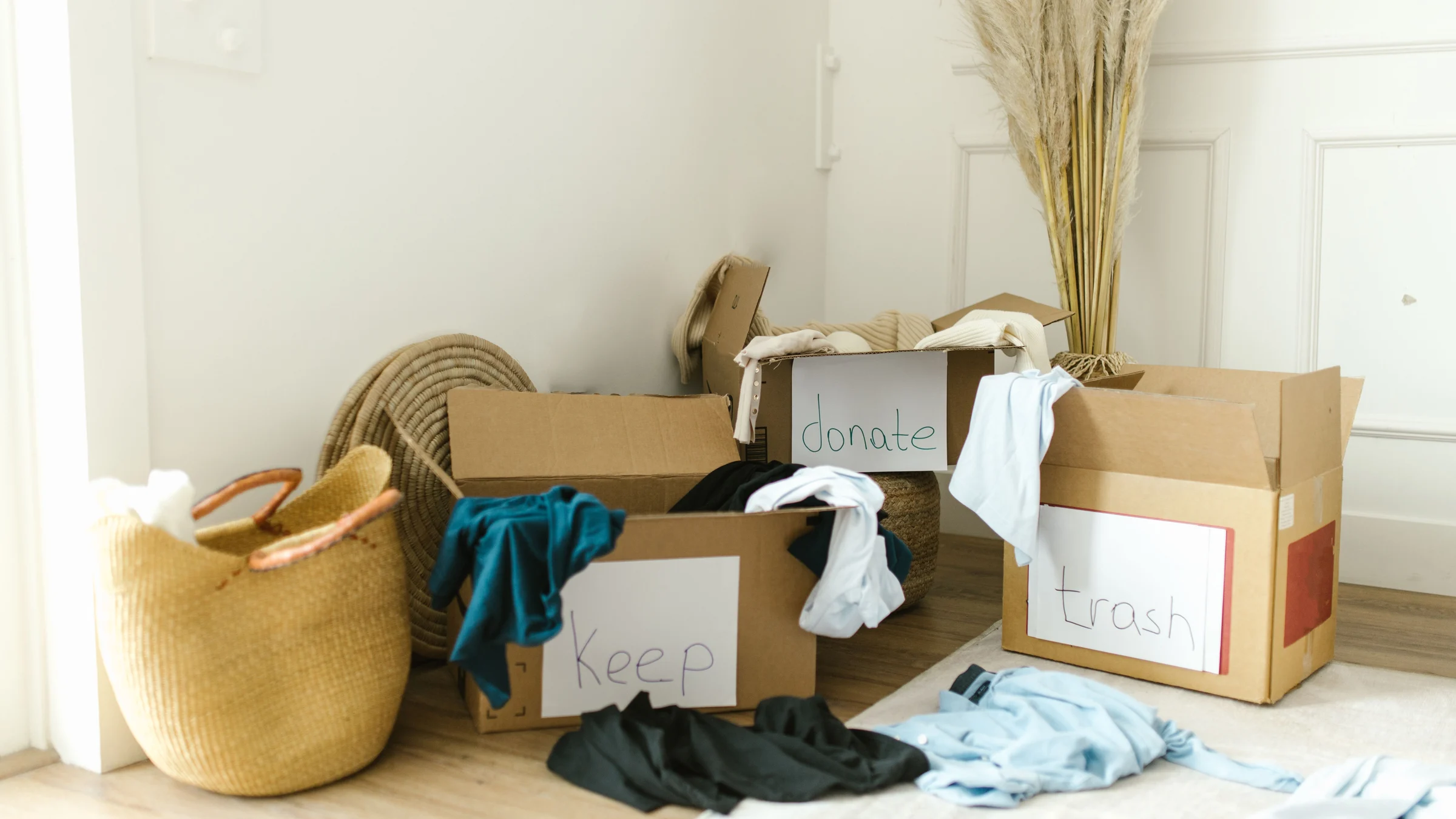 Cardboard boxes hand-labeled keep, donate and trash on a wood floor, with sorted clothes piled around them